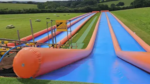 The image shows the top of four slip-and-slides situated on a green field in a rural area. They are blue plastic strips spreading out ahead and going downhill, with inflatable orange tubes on either side.