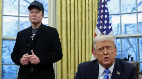 Musk standing with Trump speaking and sitting down behind a desk in front of him, in the Oval Office. 