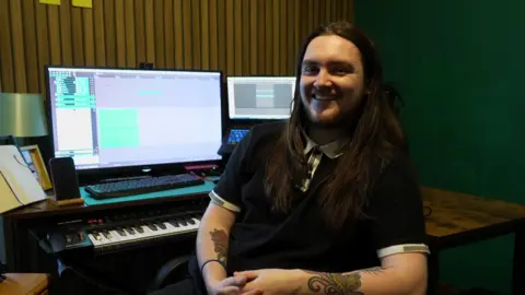 Long-haired composer Ben Randall smiles for the camera as he sits in front of two computer screens and a digital piano.