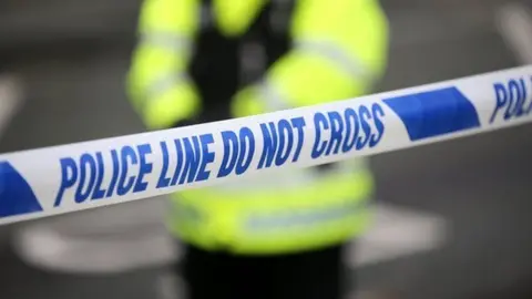 In the foreground, there is blue and white police tape that says "do not cross". In the background, out of focus, a police officer stands in their hi-vis yellow uniform and black vest, with their hands clasped in front of them.