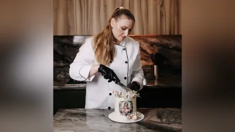 Contributed Mrs Hoian is pictured cutting a small white cake. She wears a white chef's apron and black gloves. 