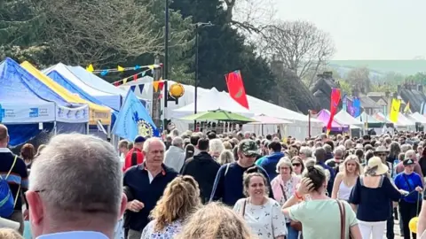 A large crowd in a Downton street, lined with gazebos, tents and flags.