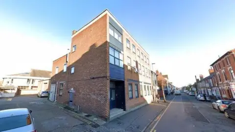 Google street view of a brick block of flats with two floors. Several terraced houses can be seen on the other side of the road with cars parked up.