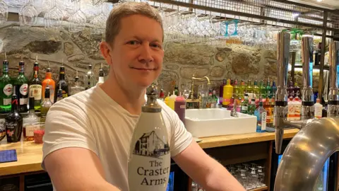 Michael Dawson is standing behind the bar in the Craster Arms and behind him are an array of spirits. In front of him is a row of beer pumps. He is smiling.
