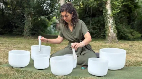 A woman with long brown curly hair, wears olive coloured robes. She is sitting cross legged on grass in front of several tress and is creating music from several white bowls in front of her of varying sizes.