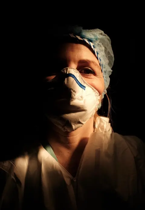 Paolo Miranda A female nurse wearing a face mask with a dark background behind her.