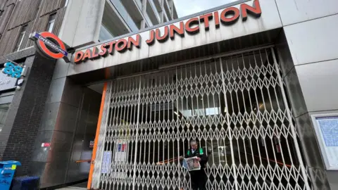 PA Media A man stands looking at a newspaper outside Dalston Junction Overground station, closed because of strike action