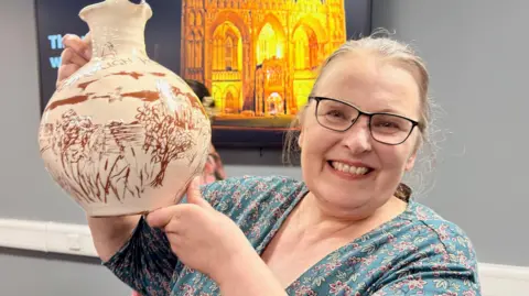 Emily Steele/Toby Wood Hilary Steele, who has glasses and tied-back blond hair, looks at the camera and smiles. She is holding up a cream-coloured jug with a decorative red pattern on it.