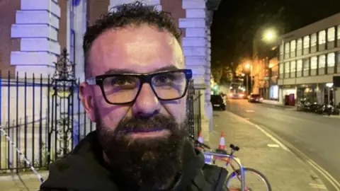 Alex Dunlop/BBC A man with dark glasses and a beard. He is standing outside in a city centre street at night. He is wearing a black coat. Behind him is a bicycle.