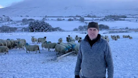 Shows a man in a grey jumper and woollen hat with a flock of sheep in the background and snow on the ground and tree tops.