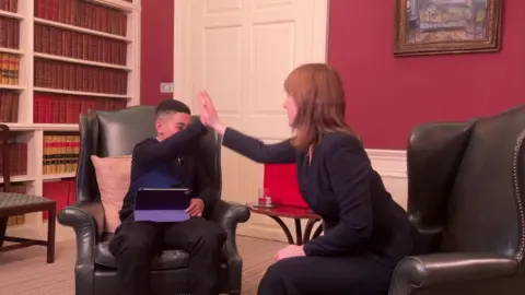 La primera ministra Rachel Reeves está sentada en una silla de cuero a la derecha de la foto, con Theo, de 11 años, sentado en una silla similar a mi lado. Ambos levantaron sus manos al cielo el uno hacia el otro.