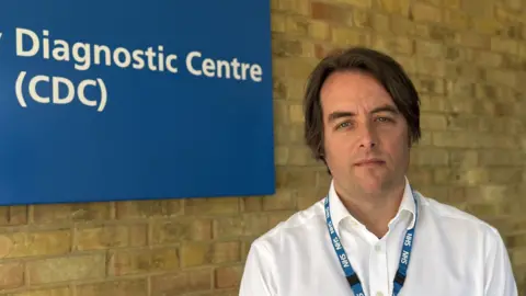 Elliot Deady/BBC A man - Thom Lafferty - looking straight at the camera. He is standing in front of a brick wall next to a blue sign. He is wearing a white shirt with a blue NHS lanyard around his neck.