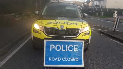 Police car behind sign reading "POLICE ROAD CLOSED"