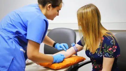 A female nurse with short dark hair, wearing a light blue uniform, leans over a female patient while wearing blue latex gloves which she is wearing on her hands as she prods the woman's right arm, which is resting on an orange cushion. The woman she is prodding has long light brown hair, which hangs over her face, and wears a dark blue top with a pattern of flowers on it.