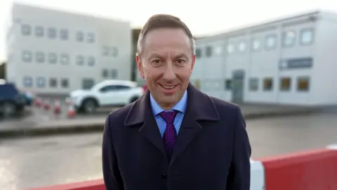 A man with brown hair, wearing a navy jacket, blue coat and purple tie. He is stood outside an out of focus white office block with cars parked behind him.