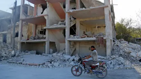 AFP A Syrian man rides a motorbike past a building reportedly damaged by an air strike in Jisr al-Shughour (4 September 2018)