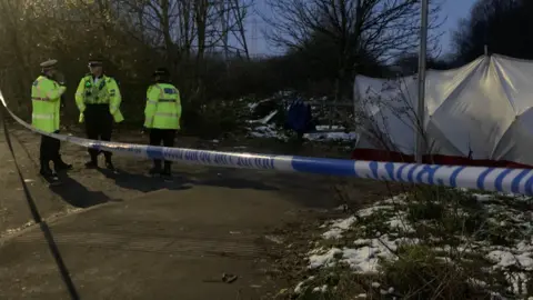 Three police officers wearing yellow hi-vis jackets stand behind tape cordoning off the scene where the baby's remains were found.