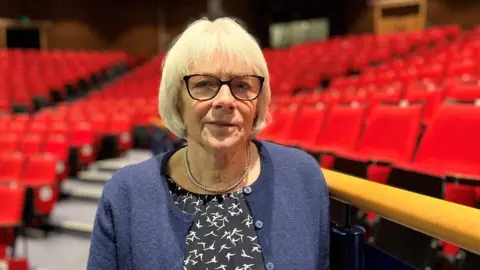 a woman with grey hair and black rimmed glasses wearing navy clothes in a theatre auditorium surrounded by red seats