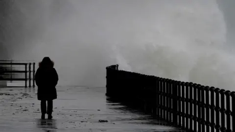A silhouetted person walks along a paved seafront wearing a raincoat. To the right of the image, sea splash from a large wave spills over a silhouetted fence on to the pavement behind the person. Conditions appear wet, windy and grey.