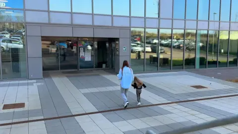 An image of the backs of Corrina and her three-year-old daughter as they make their way into the entrance of Five Wells. Corrina is wearing a large blue shirt and grey trousers.