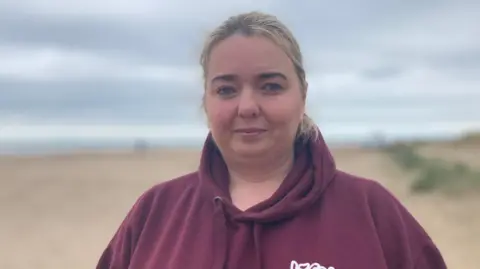 Shows a woman with fair hair in a purple hoodie standing on a beach