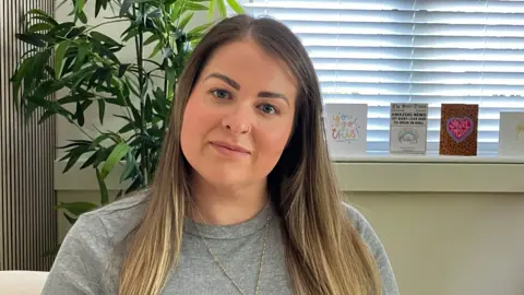 A woman looks at the camera. She is wearing a grey top and a gold chain with a pendant. She has long hair. Behind her is a plant and some cards on a window sill.