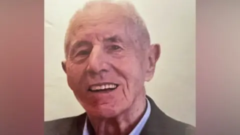An elderly man in a dark jacket and blue collared shirt.