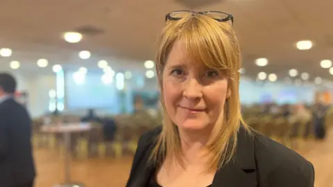 A woman with shoulder-length hair wearing a dark blazer stands in a conference or event venue. Glasses rest on the person's head. Rows of chairs and soft overhead lighting appear blurred in the background due to shallow depth of field.