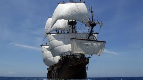 A historic wooden ship with about half a dozen large white sails billowing in the wind. The ship is on the open ocean, with no land visible on the horizon. It is a sunny day, with a clear blue sky above it.