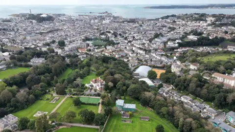 BBC A drone photo of St Helier and some of Jersey. In the distance it shows the sea and then shows buildings. There is also a large site of green as well as trees.
