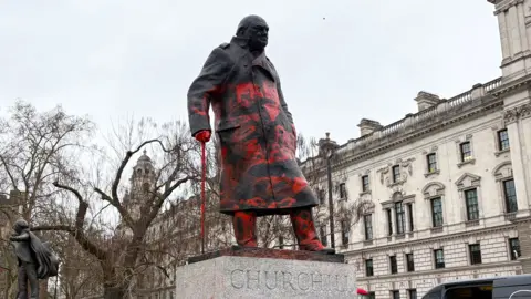 Winston Churchill statue seen from the left front side, looking up at the statue, covered in red paint. Buildings in the background 