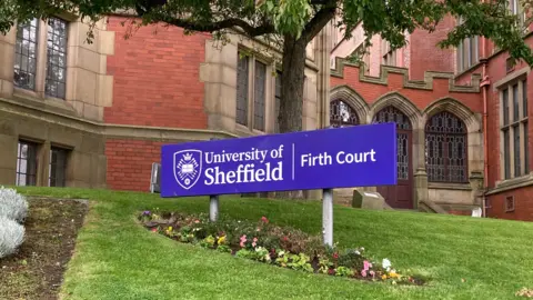A blue sign says "University of Sheffield, Firth Court" and is positioned on a grass lawn outside a red brick built university building