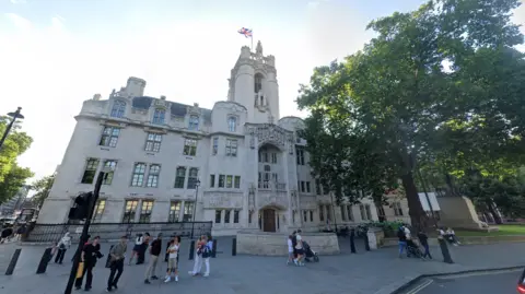 Google An image of the outside of the Supreme Court in London. It is a large white-coloured building with a tower and a Union flag flies from a mast on top. In front of the building there are people standing on the pavement. There are trees either side.