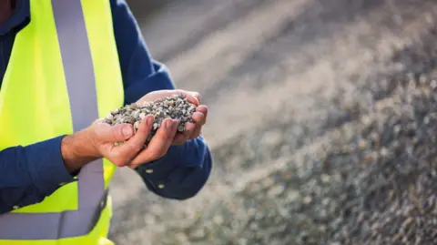 A man in a blue shirt covered with a hi-viz jacket holds up some gravel in his hands, behind him is a pile of gravel