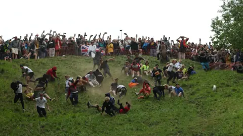 A large crowd of people falling down a steep grassy slope chasing a wheel of cheese