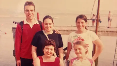 Family A woman sat down with her four children stood around her. They are at the beach in front of the sea. It is a sepia-tinted photograph.
