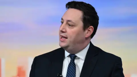 Houchen is looking to the side and is wearing a back jacket, white shirt and blue tie with white polka dots. 