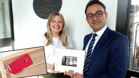 SBUHB Nicola Purdie, who has medium length blonde hair and is wearing a white t-shirt, stands next to surgeon Reza Arya, who is wearing a blue suit and tie and has short dark hair and glasses. Both are smiling and are jointly holding a framed painting of a landscape showing a promenade with people, trees and the sea in the background. There is an insert into the picture of a close up of a handwritten tag on the back of the frame, next to the words "crazy idea".
