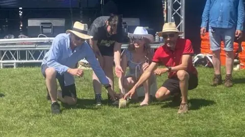 Sara Dunleavy A group of four people crouch, ready to roll a wheel of cheese. Three of them are wearing cowboy hats and two of them have their hands placed on the wheel.