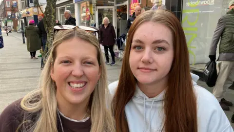 Vikki Irwin/BBC Two women smiling at the camera. The one on the left has long hair and glasses on her head and is wearing a brown jumper. The one on the right has long hair and a white jumper. They are standing in the street.