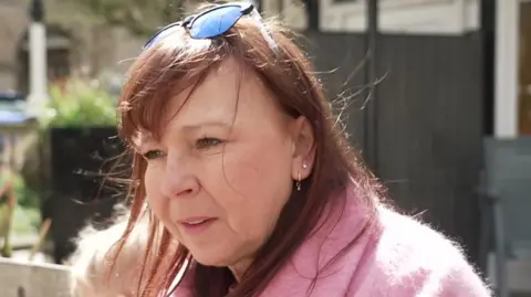 June Carson, 62, who has red hair, a pair of sunglasses resting on the top of her head and is wearing a pink, fluffy fleece, speaks to someone off camera with a concerned expression.