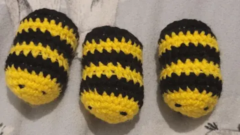 Jane Herman Three close up crocheted little bees. They are oval with yellow bodies and black lines with little black eyes.