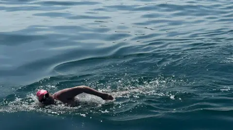 Lindsey Walke, wearing a pink swim cap and black goggles, in the sea swimming. The water around her is a navy blue. 