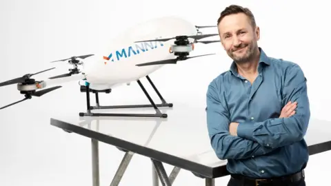 Manna Aero Bobby Healy, wearing a blue shirt and sporting a short beard, stands with his arms crossed in front of his company's drone