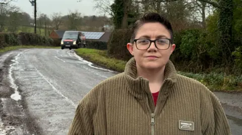Luca Straker (short hair, glasses, green zip up top) standing next to rural road lined with trees. A van is rounding the bend in the distance.