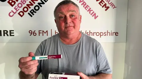 A headshot of man in a grey t-shirt, holding a red and white dispenser that is a new drug to treat asthma. He is also holding a medicine box that reads "Fasenra" in burgundy writing. 