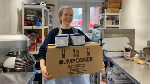 Liz is holding three boxes full of cookies in her bakery. She is wearing a hair net and an apron