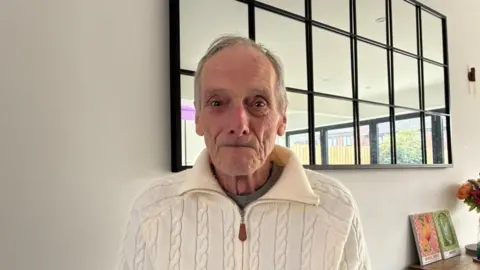 Harold Pearson stood in front of a large mirror on a white wall. He has short grey hair and wears a cream jumper with a zip down the front.