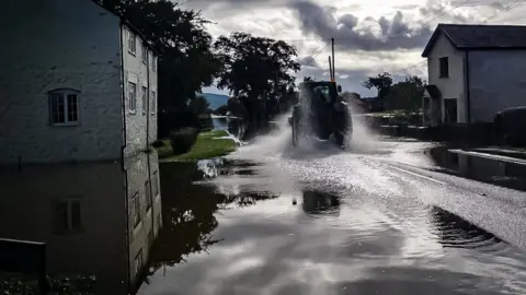 Lucy Roberts tractor is driving through water, with spray coming from is tyres, on a flooded road through a village