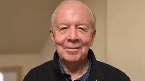 Chris Robertshaw, wo has receding short grey hair and is wearing a blue checked shirt under a black jacket.